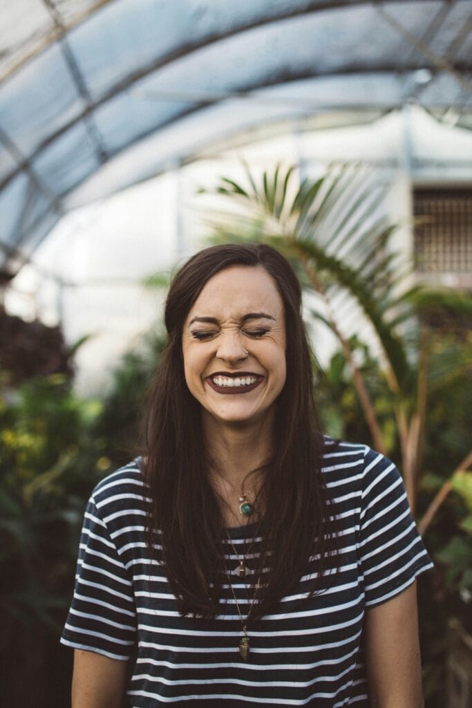 Woman with long hair smiles broadly, wearing a striped shirt, surrounded by lush greenery in a greenhouse.