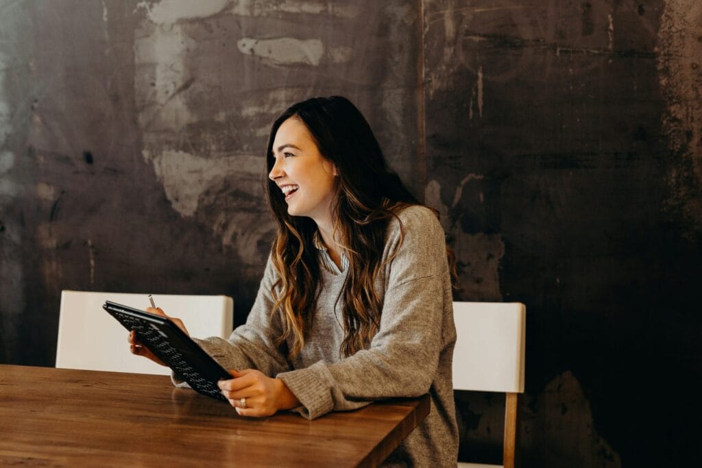 A woman with long hair laughs while holding a tablet, seated at a wooden table against a textured dark wall.