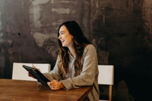 A woman with long hair laughs while holding a tablet, seated at a wooden table against a textured dark wall.